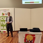 A imagem mostra Cesar Tomazini usando trajes tradicionalistas gaúchos, de pé, em frente a um banner da Secretaria da Cultura.