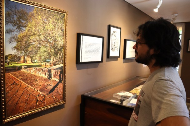 A imagem mostra um homem obserando um quadro, de moldura dourada sobre uma parede de tom marrom. O quadro é uma fotografia da região das Missões, em que se vê ima calçada antiga e ruínas avermelhadas ao fundo. O homem tem cabelo na altura dos ombros, usa óculos e barba e está de perfil.