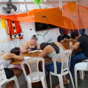 Um grupo de jovens está sentado ao redor de uma mesa grande de madeira em uma sala decorada com tecidos laranja suspensos e elementos coloridos nas paredes. Todos estão concentrados na escrita, cada um com papel e caneta, enquanto cadeiras brancas de plástico completam o espaço. À esquerda, o banner da oficina “Escrevivências” aparece parcialmente, compondo o ambiente criativo da atividade.
