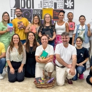 Um grupo numeroso posa para uma foto coletiva dentro de uma sala clara repleta de livros e cartazes nas paredes. As pessoas seguram cadernos, livros ou produções próprias, sugerindo o encerramento ou celebração de uma atividade literária. Algumas estão em pé e outras agachadas na frente, formando uma composição descontraída e orgulhosa dos materiais criados. No chão, um cesto com livros.
