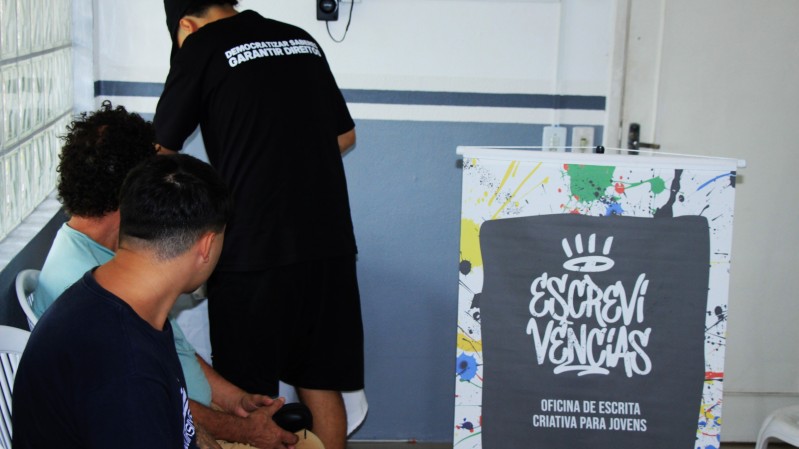 Tr&ecirc;s jovens est&atilde;o reunidos em uma sala iluminada, com cadeiras brancas de pl&aacute;stico e paredes claras. Dois deles est&atilde;o sentados lado a lado, atentos ao que ocorre &agrave; frente, enquanto outra pessoa, de p&eacute;, escreve ou organiza algo em um mural na parede. &Agrave; frente do grupo h&aacute; novamente o painel colorido da oficina &ldquo;Escreviv&ecirc;ncias&rdquo;.
