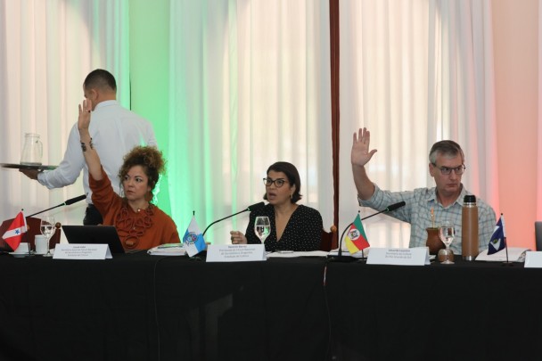 Três pessoas estão diante de uma mesa coberta com toalha preta. Duas delas estão com as mãos levantadas. 
