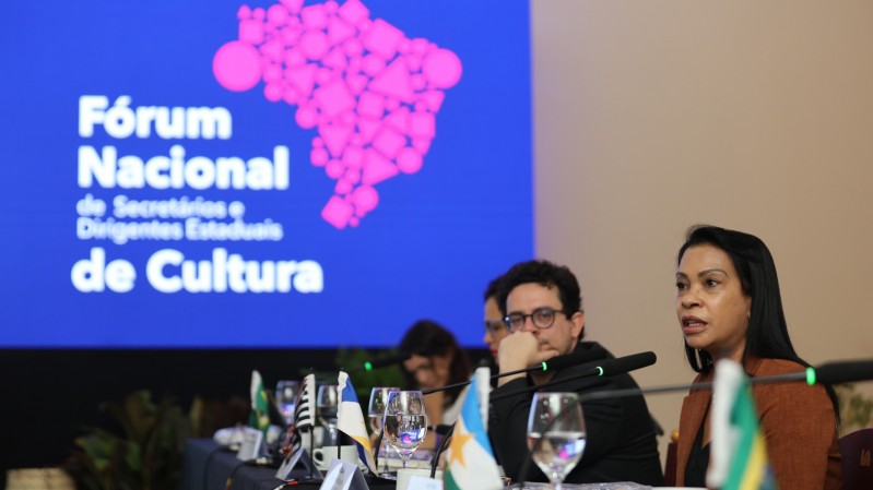 O logo do  F&oacute;rum Nacional de Secret&aacute;rios e Dirigentes Estaduais de Cultura aparece ao fundo, no canto esquerdo, em um tel&atilde;o. Na direita, aparecem quatro pessoas sentadas em frente a microfones de mesa.