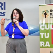 Coordenadora Regional de Educação Rosa Maria de Souza falando ao microfone. Ela está ao lado de um banner dos 400 Anos das Missões