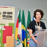 Suélen Bavaresco fala em um púlpito transparente. Atrás dela estão as bandeiras do Brasil, Rio Grande do Sul, Santo Ângelo e URI, perfiladas ao lado do banner dos 400 Anos das Missões. 