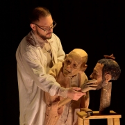 um ator em cena, vestido com figurino claro que remete a um artesão, manipula delicadamente uma marionete de aparência envelhecida. O boneco tem expressão cansada e melancólica, com traços que lembram um homem idoso, enquanto o ator ajusta suas mãos e observa atentamente o rosto da criatura. Ao lado, sobre um suporte de madeira, há outra cabeça de boneco, reforçando a ideia do processo de criação e da relação íntima entre criador e criatura, tema central do espetáculo “Geppetto”. A iluminação é quente e concentrada, destacando o trabalho manual e o clima introspectivo da cena.
