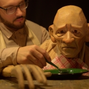 Um ator em cena, vestido com figurino claro que remete a um artesão, manipula delicadamente uma marionete de aparência envelhecida. O boneco idoso está sentado à mesa, com o olhar voltado para frente e expressão triste, enquanto o ator, ao fundo, segura uma colher e parece alimentá-lo com cuidado. A mão articulada da marionete repousa sobre a mesa, acentuando o realismo e a fragilidade do personagem. A cena transmite ternura e desconforto ao mesmo tempo, sugerindo dependência, proteção e questionamentos sobre autonomia e existência.
