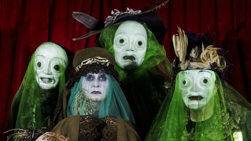 Na sexta imagem, um grupo de atrizes que comp&otilde;em o coro de &ldquo;Medea&rdquo; posa diante de um fundo avermelhado. Todas usam m&aacute;scaras claras de olhos grandes e express&otilde;es fixas, cobertas por v&eacute;us verdes transl&uacute;cidos que criam um efeito espectral. No centro, uma figura sem m&aacute;scara, com maquiagem p&aacute;lida e olhar direto, contrasta com as demais, refor&ccedil;ando a tens&atilde;o entre indiv&iacute;duo e coletivo. A imagem transmite um clima ritual&iacute;stico e perturbador, dialogando com a trag&eacute;dia, a viol&ecirc;ncia simb&oacute;lica e a dimens&atilde;o coral da encena&ccedil;&atilde;o.
