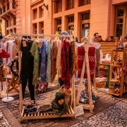 Fotografia de feira ao ar livre, realizada na Travessa dos Cataventos, mostra uma arara de madeira repleta de pe&ccedil;as artesanais de vestu&aacute;rio, como cachec&oacute;is e tecidos em cores variadas, incluindo tons vivos de vermelho, azul, verde e amarelo. A estrutura de exposi&ccedil;&atilde;o est&aacute; posicionada sobre o cal&ccedil;amento de pedras, e, abaixo dela, h&aacute; acess&oacute;rios como bolsas e um manequim com gorro e cachecol. &Agrave; esquerda, uma pessoa segura um item de roupa enquanto carrega uma bolsa de tecido com a frase &ldquo;Fazer do mundo a p&aacute;tria da humanidade&rdquo;. Outras pessoas circulam entre as barracas ao fundo, onde tamb&eacute;m s&atilde;o expostos pequenos objetos artesanais em m&oacute;veis de madeira. A ilumina&ccedil;&atilde;o natural combinada com tons quentes das constru&ccedil;&otilde;es cria uma atmosfera acolhedora e convidativa.

