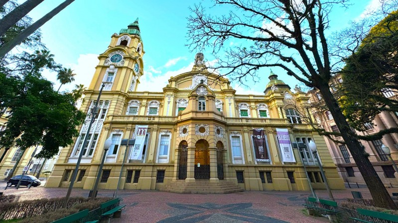 A imagem mostra um edif&iacute;cio hist&oacute;rico de arquitetura imponente, visto a partir de um &acirc;ngulo baixo que valoriza sua altura e detalhes ornamentais. A constru&ccedil;&atilde;o tem fachada em tons claros de amarelo e bege, com elementos decorativos em relevo, janelas alinhadas e uma torre com rel&oacute;gio no lado esquerdo. No topo dessa torre, destaca-se uma estrutura com cobertura em verde, que contrasta com o c&eacute;u azul intenso ao fundo.

&Agrave; frente do pr&eacute;dio, h&aacute; uma pequena pra&ccedil;a com piso de pedras e alguns bancos verdes distribu&iacute;dos ao redor. &Aacute;rvores de diferentes tipos emolduram a cena, incluindo palmeiras altas &agrave; esquerda e uma &aacute;rvore de galhos quase sem folhas &agrave; direita, cujos ramos se espalham pelo c&eacute;u. A luz do dia ilumina toda a composi&ccedil;&atilde;o, refor&ccedil;ando as cores vibrantes e os detalhes da arquitetura. Pr&oacute;ximo &agrave; entrada central do edif&iacute;cio, &eacute; poss&iacute;vel ver banners verticais pendurados, com textos e s&iacute;mbolos que indicam exposi&ccedil;&otilde;es ou atividades culturais no local.