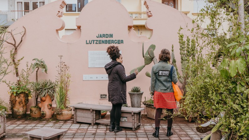Fotografia de duas pessoas em frente a um muro decorativo de cor clara com formas arredondadas e o nome &ldquo;Jardim Lutzenberger&rdquo; em letras verdes ao centro. O espa&ccedil;o &eacute; um jardim, cercado por diferentes tipos de plantas, incluindo cactos e vasos com esp&eacute;cies variadas dispostos ao redor. Uma das pessoas gesticula com a m&atilde;o enquanto conversa, e ambas est&atilde;o voltadas para o local, pr&oacute;ximas a um banco de madeira baixo. Ao fundo, percebe-se a fachada de um pr&eacute;dio residencial com janelas e sacadas, compondo um ambiente urbano.
