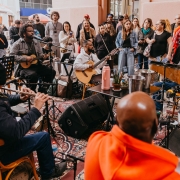 A imagem retrata uma apresenta&ccedil;&atilde;o musical realizada em um espa&ccedil;o interno, com diversos m&uacute;sicos reunidos em c&iacute;rculo e um p&uacute;blico observando ao redor. O ambiente tem apar&ecirc;ncia descontra&iacute;da e acolhedora, semelhante a um caf&eacute;, centro cultural ou sala de eventos. Em primeiro plano, aparecem m&uacute;sicos tocando diferentes instrumentos. &Agrave; esquerda, uma pessoa toca uma flauta transversal. No centro, dois m&uacute;sicos sentados executam instrumentos de cordas, um deles com um viol&atilde;o ac&uacute;stico. &Agrave; direita, outro participante est&aacute; cercado por instrumentos de percuss&atilde;o, incluindo sinos e pequenos tambores. Microfones, caixas de som, suportes para partituras e cabos espalhados pelo ch&atilde;o indicam uma apresenta&ccedil;&atilde;o ao vivo. Ao redor dos artistas, um grande grupo de espectadores acompanha atentamente a performance. Algumas pessoas est&atilde;o em p&eacute;, enquanto outras permanecem sentadas. 
