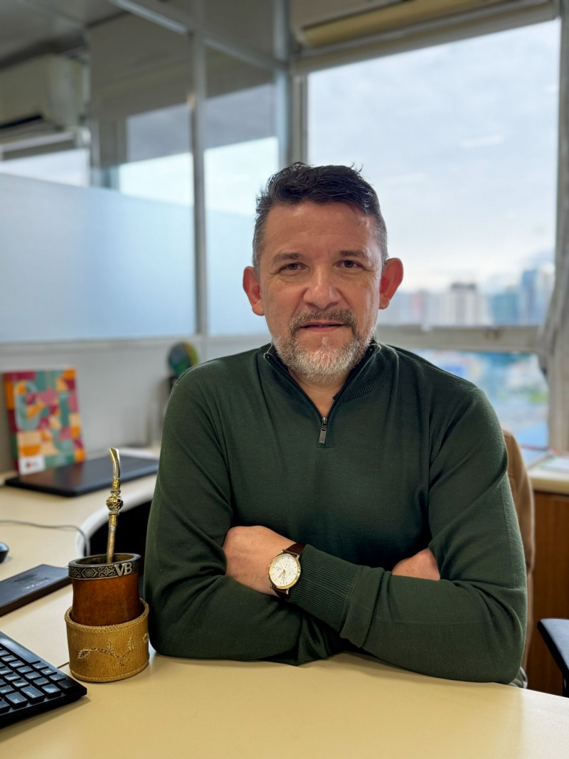 Fotografia de um homem posando sentado atr&aacute;s de uma mesa em um escrit&oacute;rio, com os bra&ccedil;os apoiados &agrave; frente do corpo. A pessoa usa uma blusa de cor escura com gola alta e um rel&oacute;gio no pulso. Sobre a mesa, h&aacute; um teclado e um recipiente com uma cuia e uma bomba de chimarr&atilde;o, al&eacute;m de um caderno ou livro com capa colorida ao lado. Ao fundo, uma grande janela revela a vista de pr&eacute;dios urbanos sob o c&eacute;u parcialmente nublado, com aparelhos de ar-condicionado instalados acima da janela e divis&oacute;rias de vidro compondo o ambiente.

