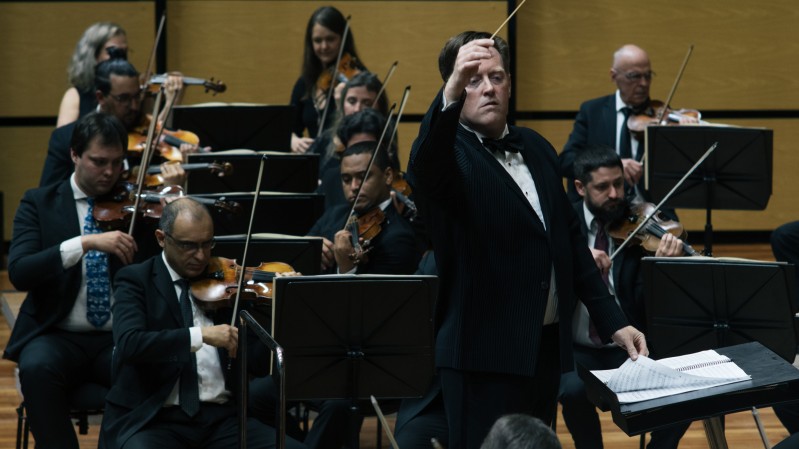 A imagem mostra uma apresenta&ccedil;&atilde;o de uma orquestra sinf&ocirc;nica em um palco de concertos. O fundo &eacute; formado por pain&eacute;is de madeira em tom claro, caracter&iacute;sticos de salas de espet&aacute;culo com tratamento ac&uacute;stico. No centro da cena est&aacute; o maestro, em p&eacute;, usando fraque preto, camisa branca e gravata-borboleta. Ele est&aacute; voltado para a orquestra, com o bra&ccedil;o direito elevado e a m&atilde;o aberta, conduzindo os m&uacute;sicos durante a execu&ccedil;&atilde;o da obra. &Agrave; sua frente h&aacute; um p&uacute;lpito com a partitura aberta. Ao redor do maestro est&atilde;o diversos integrantes da orquestra, vestidos com trajes formais pretos. Em primeiro plano e nas laterais aparecem violinistas e violistas sentados, segurando seus instrumentos e arcos em posi&ccedil;&atilde;o de execu&ccedil;&atilde;o, concentrados na leitura das partituras apoiadas em estantes pretas. Ao fundo, outros m&uacute;sicos tamb&eacute;m tocam instrumentos de cordas, compondo diferentes fileiras da orquestra.