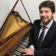 A imagem mostra o pianista Fernando Rauber, um homem posicionado ao lado de um cravo histórico, em um ambiente interno de paredes brancas. O instrumento ocupa a maior parte do lado esquerdo da fotografia. Sua tampa está aberta, revelando o interior com as cordas e a estrutura de madeira. O móvel apresenta acabamento em madeira escura com detalhes ornamentais, e o teclado, com teclas claras e escuras, está em primeiro plano. À direita está o homem, sentado ou apoiado próximo ao teclado do instrumento. Ele usa terno escuro, camisa branca e gravata listrada em tons de azul, vermelho e branco. Tem cabelos castanhos curtos, barba e bigode aparados, e sorri diretamente para a câmera. Suas mãos repousam sobre o teclado do cravo, em uma posição que sugere que está prestes a tocar ou demonstrar o instrumento.