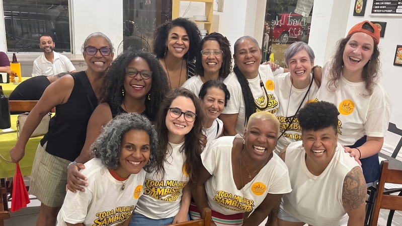 A imagem mostra um grupo de doze mulheres reunidas para uma fotografia em um ambiente interno, provavelmente um caf&eacute;, restaurante ou espa&ccedil;o de conviv&ecirc;ncia. Todas est&atilde;o sorrindo e voltadas para a c&acirc;mera, transmitindo um clima de descontra&ccedil;&atilde;o, acolhimento e celebra&ccedil;&atilde;o. As participantes est&atilde;o distribu&iacute;das em duas fileiras. Na frente, cinco mulheres aparecem sentadas ou agachadas, enquanto as demais est&atilde;o em p&eacute; logo atr&aacute;s, com algumas apoiando as m&atilde;os nos ombros ou abra&ccedil;ando as colegas. A maioria veste camisetas brancas estampadas com a frase "Toda Mulher &eacute; Sambista" em letras amarelas e pretas. Algumas tamb&eacute;m usam adesivos circulares amarelos presos &agrave; roupa. Outras participantes usam blusas pretas ou brancas lisas. As mulheres apresentam diferentes caracter&iacute;sticas f&iacute;sicas, como variados tons de pele, tipos e cores de cabelo &mdash; incluindo cabelos cacheados, crespos, lisos, grisalhos e curtos. Algumas usam &oacute;culos, e uma pessoa, &agrave; direita da imagem, usa um bon&eacute; laranja virado para tr&aacute;s. No canto inferior direito, uma participante tem uma tatuagem vis&iacute;vel no bra&ccedil;o esquerdo. Ao fundo, h&aacute; mesas, cadeiras e grandes janelas de vidro. No lado esquerdo aparece um homem sentado, sorrindo, ao fundo do ambiente. Pela janela &eacute; poss&iacute;vel ver parte da rua e um ve&iacute;culo vermelho estacionado do lado de fora.