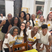A imagem mostra um grupo de doze mulheres reunidas para uma fotografia em um ambiente interno, provavelmente um caf&eacute;, restaurante ou espa&ccedil;o de conviv&ecirc;ncia. Todas est&atilde;o sorrindo e voltadas para a c&acirc;mera, transmitindo um clima de descontra&ccedil;&atilde;o, acolhimento e celebra&ccedil;&atilde;o. As participantes est&atilde;o distribu&iacute;das em duas fileiras. Na frente, cinco mulheres aparecem sentadas ou agachadas, enquanto as demais est&atilde;o em p&eacute; logo atr&aacute;s, com algumas apoiando as m&atilde;os nos ombros ou abra&ccedil;ando as colegas. A maioria veste camisetas brancas estampadas com a frase "Toda Mulher &eacute; Sambista" em letras amarelas e pretas. Algumas tamb&eacute;m usam adesivos circulares amarelos presos &agrave; roupa. Outras participantes usam blusas pretas ou brancas lisas. As mulheres apresentam diferentes caracter&iacute;sticas f&iacute;sicas, como variados tons de pele, tipos e cores de cabelo &mdash; incluindo cabelos cacheados, crespos, lisos, grisalhos e curtos. Algumas usam &oacute;culos, e uma pessoa, &agrave; direita da imagem, usa um bon&eacute; laranja virado para tr&aacute;s. No canto inferior direito, uma participante tem uma tatuagem vis&iacute;vel no bra&ccedil;o esquerdo. Ao fundo, h&aacute; mesas, cadeiras e grandes janelas de vidro. No lado esquerdo aparece um homem sentado, sorrindo, ao fundo do ambiente. Pela janela &eacute; poss&iacute;vel ver parte da rua e um ve&iacute;culo vermelho estacionado do lado de fora.