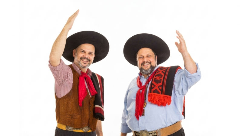 A imagem mostra duas pessoas lado a lado, em p&eacute;, diante de um fundo branco liso. Ambas sorriem para a c&acirc;mera e fazem um gesto de cumprimento com um dos bra&ccedil;os erguidos, como se estivessem acenando. &Agrave; esquerda, a pessoa veste um chap&eacute;u ga&uacute;cho preto de aba larga, camisa xadrez em tons de vermelho e branco com as mangas dobradas, colete marrom e um len&ccedil;o vermelho amarrado ao pesco&ccedil;o. Sobre o ombro esquerdo, h&aacute; uma faixa ou pala preta com detalhes em vermelho. &Agrave; direita, a pessoa tamb&eacute;m usa um chap&eacute;u ga&uacute;cho preto de aba larga, camisa azul-clara de mangas compridas e um len&ccedil;o vermelho no pesco&ccedil;o. Sobre o ombro esquerdo, h&aacute; uma pala preta com desenhos geom&eacute;tricos vermelhos e franjas vermelhas na extremidade. As duas pessoas ocupam a regi&atilde;o central da imagem, est&atilde;o voltadas para a frente e demonstram uma express&atilde;o alegre e acolhedora.