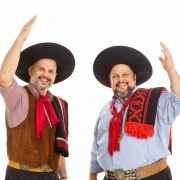 A imagem mostra duas pessoas lado a lado, em p&eacute;, diante de um fundo branco liso. Ambas sorriem para a c&acirc;mera e fazem um gesto de cumprimento com um dos bra&ccedil;os erguidos, como se estivessem acenando. &Agrave; esquerda, a pessoa veste um chap&eacute;u ga&uacute;cho preto de aba larga, camisa xadrez em tons de vermelho e branco com as mangas dobradas, colete marrom e um len&ccedil;o vermelho amarrado ao pesco&ccedil;o. Sobre o ombro esquerdo, h&aacute; uma faixa ou pala preta com detalhes em vermelho. &Agrave; direita, a pessoa tamb&eacute;m usa um chap&eacute;u ga&uacute;cho preto de aba larga, camisa azul-clara de mangas compridas e um len&ccedil;o vermelho no pesco&ccedil;o. Sobre o ombro esquerdo, h&aacute; uma pala preta com desenhos geom&eacute;tricos vermelhos e franjas vermelhas na extremidade. As duas pessoas ocupam a regi&atilde;o central da imagem, est&atilde;o voltadas para a frente e demonstram uma express&atilde;o alegre e acolhedora.
