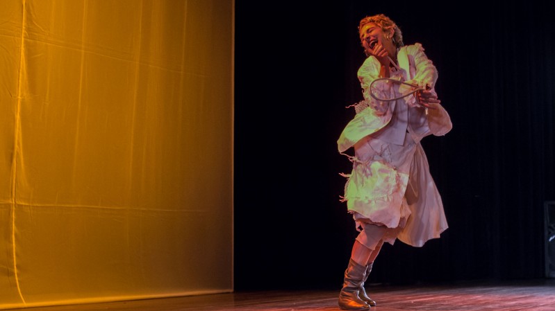 A imagem mostra uma cena de espet&aacute;culo teatral em um palco. O fundo &eacute; dividido em duas &aacute;reas: &agrave; esquerda h&aacute; um grande painel iluminado por uma luz &acirc;mbar intensa, que cria uma atmosfera quente, enquanto &agrave; direita o fundo &eacute; escuro, destacando a artista em cena. O piso de madeira do palco aparece na parte inferior da imagem. &Agrave; direita, uma artista est&aacute; em movimento, com o corpo inclinado para tr&aacute;s e uma das pernas cruzada &agrave; frente da outra, sugerindo um giro ou um passo de dan&ccedil;a. Ela sorri amplamente, com a cabe&ccedil;a levemente inclinada para tr&aacute;s, transmitindo expressividade e alegria. Usa um vestido branco de mangas longas e volumosas, com camadas de tecido que se movimentam junto ao corpo. Cal&ccedil;a botas de cano m&eacute;dio em tom prateado ou met&aacute;lico. A performer segura um pequeno objeto circular com aro e fios entrela&ccedil;ados, semelhante a um bastidor ou instrumento c&ecirc;nico, junto ao peito. A ilumina&ccedil;&atilde;o do palco projeta tons dourados e esverdeados sobre sua roupa, criando contrastes de cor e refor&ccedil;ando a sensa&ccedil;&atilde;o de movimento. A composi&ccedil;&atilde;o enfatiza a presen&ccedil;a da artista e o dinamismo da apresenta&ccedil;&atilde;o, com o amplo espa&ccedil;o vazio &agrave; esquerda equilibrando visualmente a cena.