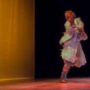 A imagem mostra uma cena de espet&aacute;culo teatral em um palco. O fundo &eacute; dividido em duas &aacute;reas: &agrave; esquerda h&aacute; um grande painel iluminado por uma luz &acirc;mbar intensa, que cria uma atmosfera quente, enquanto &agrave; direita o fundo &eacute; escuro, destacando a artista em cena. O piso de madeira do palco aparece na parte inferior da imagem. &Agrave; direita, uma artista est&aacute; em movimento, com o corpo inclinado para tr&aacute;s e uma das pernas cruzada &agrave; frente da outra, sugerindo um giro ou um passo de dan&ccedil;a. Ela sorri amplamente, com a cabe&ccedil;a levemente inclinada para tr&aacute;s, transmitindo expressividade e alegria. Usa um vestido branco de mangas longas e volumosas, com camadas de tecido que se movimentam junto ao corpo. Cal&ccedil;a botas de cano m&eacute;dio em tom prateado ou met&aacute;lico. A performer segura um pequeno objeto circular com aro e fios entrela&ccedil;ados, semelhante a um bastidor ou instrumento c&ecirc;nico, junto ao peito. A ilumina&ccedil;&atilde;o do palco projeta tons dourados e esverdeados sobre sua roupa, criando contrastes de cor e refor&ccedil;ando a sensa&ccedil;&atilde;o de movimento. A composi&ccedil;&atilde;o enfatiza a presen&ccedil;a da artista e o dinamismo da apresenta&ccedil;&atilde;o, com o amplo espa&ccedil;o vazio &agrave; esquerda equilibrando visualmente a cena.
