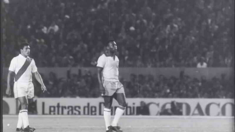 Fotograma em preto e branco de uma partida de futebol mostra Pel&eacute; e outro jogador uniformizados em campo. Pel&eacute; ocupa o centro da imagem, enquanto o outro jogador aparece &agrave; esquerda usando uma camisa com faixa diagonal. Ao fundo, arquibancadas lotadas cercam o est&aacute;dio e h&aacute; uma placa publicit&aacute;ria parcialmente vis&iacute;vel. A cena possui granula&ccedil;&atilde;o e marcas t&iacute;picas de registros hist&oacute;ricos em filme.
