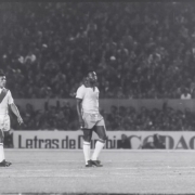 Fotograma em preto e branco de uma partida de futebol mostra Pel&eacute; e outro jogador uniformizados em campo. Pel&eacute; ocupa o centro da imagem, enquanto o outro jogador aparece &agrave; esquerda usando uma camisa com faixa diagonal. Ao fundo, arquibancadas lotadas cercam o est&aacute;dio e h&aacute; uma placa publicit&aacute;ria parcialmente vis&iacute;vel. A cena possui granula&ccedil;&atilde;o e marcas t&iacute;picas de registros hist&oacute;ricos em filme.