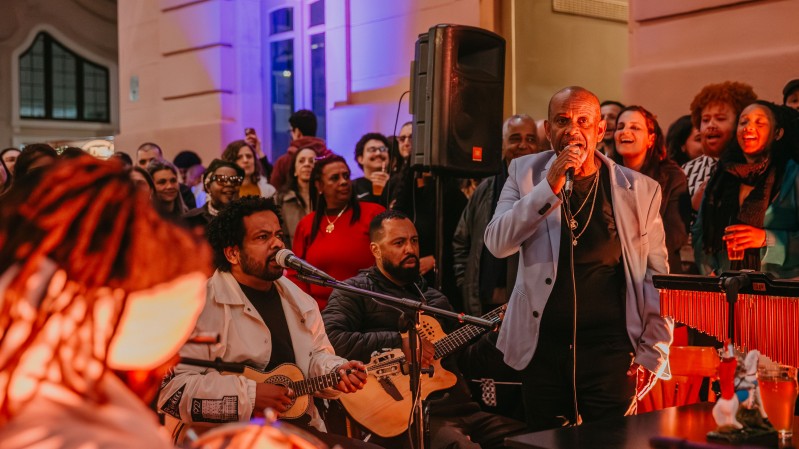 A imagem mostra uma apresenta&ccedil;&atilde;o musical ao ar livre, realizada &agrave; noite, diante da fachada iluminada de um pr&eacute;dio de arquitetura cl&aacute;ssica. A cena &eacute; marcada por uma ilumina&ccedil;&atilde;o quente, em tons alaranjados e avermelhados, que destaca os artistas em primeiro plano, enquanto luzes azuladas iluminam o edif&iacute;cio ao fundo. No centro da imagem, um homem veste blazer cinza-claro sobre camiseta preta e segura um microfone com uma das m&atilde;os, aparentando cantar ou falar ao p&uacute;blico. Ao seu lado esquerdo, um m&uacute;sico sentado canta em um microfone enquanto toca um cavaquinho. Ele usa uma jaqueta clara e est&aacute; concentrado na apresenta&ccedil;&atilde;o. Logo atr&aacute;s dele, outro m&uacute;sico, tamb&eacute;m sentado, toca um viol&atilde;o ac&uacute;stico. &Agrave; direita da cena, aparece parte de um instrumento de percuss&atilde;o com barras vermelhas, e uma pessoa posicionada atr&aacute;s dele, parcialmente vis&iacute;vel. Em primeiro plano, no canto inferior esquerdo, v&ecirc;-se desfocada a parte de tr&aacute;s da cabe&ccedil;a de outra pessoa, com cabelos em tran&ccedil;as, sugerindo a presen&ccedil;a de mais m&uacute;sicos pr&oacute;ximos &agrave; c&acirc;mera. Ao fundo, um grande p&uacute;blico acompanha a apresenta&ccedil;&atilde;o. As pessoas est&atilde;o reunidas em p&eacute;, sorrindo, algumas cantando, conversando ou registrando o momento com celulares. Um alto-falante de grande porte est&aacute; instalado atr&aacute;s dos m&uacute;sicos, refor&ccedil;ando o car&aacute;ter de show ou apresenta&ccedil;&atilde;o p&uacute;blica. 