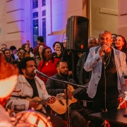A imagem mostra uma apresenta&ccedil;&atilde;o musical ao ar livre, realizada &agrave; noite, diante da fachada iluminada de um pr&eacute;dio de arquitetura cl&aacute;ssica. A cena &eacute; marcada por uma ilumina&ccedil;&atilde;o quente, em tons alaranjados e avermelhados, que destaca os artistas em primeiro plano, enquanto luzes azuladas iluminam o edif&iacute;cio ao fundo. No centro da imagem, um homem veste blazer cinza-claro sobre camiseta preta e segura um microfone com uma das m&atilde;os, aparentando cantar ou falar ao p&uacute;blico. Ao seu lado esquerdo, um m&uacute;sico sentado canta em um microfone enquanto toca um cavaquinho. Ele usa uma jaqueta clara e est&aacute; concentrado na apresenta&ccedil;&atilde;o. Logo atr&aacute;s dele, outro m&uacute;sico, tamb&eacute;m sentado, toca um viol&atilde;o ac&uacute;stico. &Agrave; direita da cena, aparece parte de um instrumento de percuss&atilde;o com barras vermelhas, e uma pessoa posicionada atr&aacute;s dele, parcialmente vis&iacute;vel. Em primeiro plano, no canto inferior esquerdo, v&ecirc;-se desfocada a parte de tr&aacute;s da cabe&ccedil;a de outra pessoa, com cabelos em tran&ccedil;as, sugerindo a presen&ccedil;a de mais m&uacute;sicos pr&oacute;ximos &agrave; c&acirc;mera. Ao fundo, um grande p&uacute;blico acompanha a apresenta&ccedil;&atilde;o. As pessoas est&atilde;o reunidas em p&eacute;, sorrindo, algumas cantando, conversando ou registrando o momento com celulares. Um alto-falante de grande porte est&aacute; instalado atr&aacute;s dos m&uacute;sicos, refor&ccedil;ando o car&aacute;ter de show ou apresenta&ccedil;&atilde;o p&uacute;blica. 