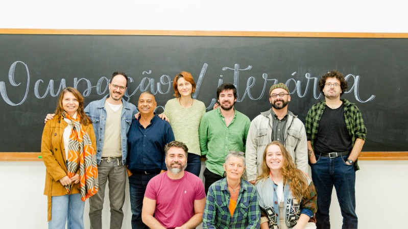 A imagem mostra um grupo de dez pessoas posando para uma fotografia em frente a um quadro negro de sala de aula. Ao fundo, ocupando toda a largura da imagem, h&aacute; um quadro de giz escuro onde est&aacute; escrita, em letras grandes e cursivas, a frase "Improvisa&ccedil;&atilde;o Liter&aacute;ria". A parede abaixo do quadro &eacute; branca. As pessoas est&atilde;o distribu&iacute;das em duas fileiras. Na fileira de tr&aacute;s, sete pessoas permanecem em p&eacute;, lado a lado. Da esquerda para a direita, h&aacute; uma mulher com cabelos longos ruivos, usando casaco mostarda e um len&ccedil;o estampado em tons de laranja, branco e cinza; um homem de barba curta, &oacute;culos e blazer azul-claro sobre camiseta bege; um homem de baixa estatura com camisa azul-marinho; uma mulher de cabelos ruivos presos, vestindo blusa verde-clara; um homem de barba curta com camisa verde de mangas dobradas; um homem de barba, &oacute;culos, bon&eacute; e jaqueta bege sobre camiseta escura; e um homem de cabelos cacheados, &oacute;culos, camiseta preta e camisa xadrez verde aberta. Na fileira da frente, tr&ecirc;s pessoas est&atilde;o agachadas ou sentadas. &Agrave; esquerda, um homem de barba grisalha veste camiseta rosa; ao centro, uma pessoa de cabelos curtos grisalhos usa camisa xadrez em tons de verde e azul sobre camiseta laranja; &agrave; direita, uma mulher de cabelos longos ruivos sorri para a c&acirc;mera, usando camisa azul-clara e um casaco claro apoiado sobre os ombros. Todos est&atilde;o voltados para a c&acirc;mera, com express&otilde;es sorridentes ou descontra&iacute;das.