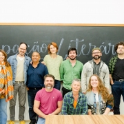 A imagem mostra um grupo de dez pessoas posando para uma fotografia em frente a um quadro negro de sala de aula. Ao fundo, ocupando toda a largura da imagem, h&aacute; um quadro de giz escuro onde est&aacute; escrita, em letras grandes e cursivas, a frase "Improvisa&ccedil;&atilde;o Liter&aacute;ria". A parede abaixo do quadro &eacute; branca. As pessoas est&atilde;o distribu&iacute;das em duas fileiras. Na fileira de tr&aacute;s, sete pessoas permanecem em p&eacute;, lado a lado. Da esquerda para a direita, h&aacute; uma mulher com cabelos longos ruivos, usando casaco mostarda e um len&ccedil;o estampado em tons de laranja, branco e cinza; um homem de barba curta, &oacute;culos e blazer azul-claro sobre camiseta bege; um homem de baixa estatura com camisa azul-marinho; uma mulher de cabelos ruivos presos, vestindo blusa verde-clara; um homem de barba curta com camisa verde de mangas dobradas; um homem de barba, &oacute;culos, bon&eacute; e jaqueta bege sobre camiseta escura; e um homem de cabelos cacheados, &oacute;culos, camiseta preta e camisa xadrez verde aberta. Na fileira da frente, tr&ecirc;s pessoas est&atilde;o agachadas ou sentadas. &Agrave; esquerda, um homem de barba grisalha veste camiseta rosa; ao centro, uma pessoa de cabelos curtos grisalhos usa camisa xadrez em tons de verde e azul sobre camiseta laranja; &agrave; direita, uma mulher de cabelos longos ruivos sorri para a c&acirc;mera, usando camisa azul-clara e um casaco claro apoiado sobre os ombros. Todos est&atilde;o voltados para a c&acirc;mera, com express&otilde;es sorridentes ou descontra&iacute;das.