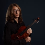 Retrato de estúdio de uma pessoa sentada, segurando um violino e o arco diante de um fundo escuro em tons azulados. A pessoa veste roupa preta e usa óculos de armação fina. Os cabelos compridos e ondulados caem até a altura dos ombros. O violino, em madeira de tonalidade marrom-avermelhada, está apoiado junto ao corpo e é segurado com ambas as mãos. A iluminação direcionada destaca o instrumento e a figura retratada, criando contraste com o fundo escuro e reforçando o caráter artístico da composição.