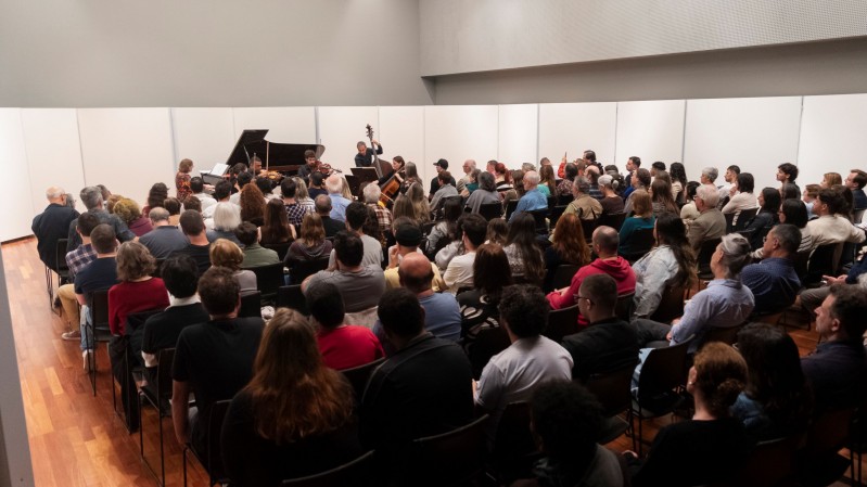 Fotografia de uma apresenta&ccedil;&atilde;o de m&uacute;sica de c&acirc;mara em uma sala de concertos de porte intimista. O p&uacute;blico ocupa todas as cadeiras do espa&ccedil;o e acompanha a apresenta&ccedil;&atilde;o voltado para um pequeno palco &agrave; frente. Entre os m&uacute;sicos s&atilde;o vis&iacute;veis um piano de cauda, instrumentos de cordas e estantes de partitura. O ambiente possui paredes claras, ilumina&ccedil;&atilde;o suave e piso de madeira, criando uma atmosfera acolhedora para a apresenta&ccedil;&atilde;o musical.