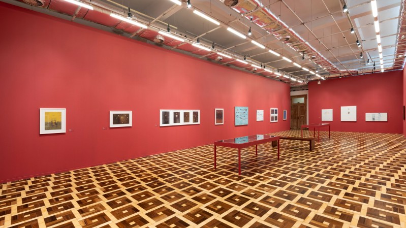 A imagem mostra uma ampla sala expositiva do MARGS, vista em perspectiva a partir de um dos cantos do ambiente. O espa&ccedil;o possui paredes pintadas em vermelho escuro, que ocupam toda a extens&atilde;o da sala. Distribu&iacute;das ao longo dessas paredes est&atilde;o diversas obras de arte emolduradas, de diferentes tamanhos, alinhadas em uma &uacute;nica altura. Entre elas h&aacute; fotografias, gravuras, desenhos e alguns trabalhos com fundo azul-claro e branco. Pequenas legendas de identifica&ccedil;&atilde;o acompanham algumas das obras. No centro da sala h&aacute; duas mesas retangulares de estrutura met&aacute;lica vermelha e tampo de vidro ou acr&iacute;lico transparente, utilizadas para expor documentos, livros ou objetos. As mesas est&atilde;o posicionadas paralelamente, deixando amplos corredores para circula&ccedil;&atilde;o. O piso &eacute; um dos elementos de maior destaque da imagem, formado por um padr&atilde;o geom&eacute;trico em madeira, com quadrados em diferentes tonalidades de marrom e bege que se repetem por toda a superf&iacute;cie. O teto &eacute; branco, com estrutura aparente, trilhos eletrificados e diversos refletores direcionados &agrave;s obras, proporcionando ilumina&ccedil;&atilde;o uniforme ao ambiente. Ao fundo, &agrave; direita, h&aacute; uma abertura que conduz a outro espa&ccedil;o da exposi&ccedil;&atilde;o. A sala est&aacute; vazia, sem visitantes.