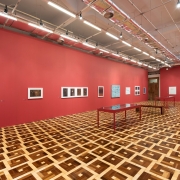 A imagem mostra uma ampla sala expositiva do MARGS, vista em perspectiva a partir de um dos cantos do ambiente. O espa&ccedil;o possui paredes pintadas em vermelho escuro, que ocupam toda a extens&atilde;o da sala. Distribu&iacute;das ao longo dessas paredes est&atilde;o diversas obras de arte emolduradas, de diferentes tamanhos, alinhadas em uma &uacute;nica altura. Entre elas h&aacute; fotografias, gravuras, desenhos e alguns trabalhos com fundo azul-claro e branco. Pequenas legendas de identifica&ccedil;&atilde;o acompanham algumas das obras. No centro da sala h&aacute; duas mesas retangulares de estrutura met&aacute;lica vermelha e tampo de vidro ou acr&iacute;lico transparente, utilizadas para expor documentos, livros ou objetos. As mesas est&atilde;o posicionadas paralelamente, deixando amplos corredores para circula&ccedil;&atilde;o. O piso &eacute; um dos elementos de maior destaque da imagem, formado por um padr&atilde;o geom&eacute;trico em madeira, com quadrados em diferentes tonalidades de marrom e bege que se repetem por toda a superf&iacute;cie. O teto &eacute; branco, com estrutura aparente, trilhos eletrificados e diversos refletores direcionados &agrave;s obras, proporcionando ilumina&ccedil;&atilde;o uniforme ao ambiente. Ao fundo, &agrave; direita, h&aacute; uma abertura que conduz a outro espa&ccedil;o da exposi&ccedil;&atilde;o. A sala est&aacute; vazia, sem visitantes.