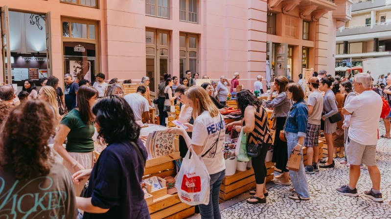 Uma feira ao ar livre acontece em frente a um edif&iacute;cio hist&oacute;rico de fachada rosa e grandes janelas. Diversas pessoas circulam entre bancas de madeira organizadas sobre a cal&ccedil;ada revestida por pedras em padr&atilde;o mosaico. Os expositores apresentam ilustra&ccedil;&otilde;es, objetos artesanais e outros produtos culturais. O movimento intenso de visitantes observando os itens e conversando entre si destaca o car&aacute;ter coletivo e cultural do evento.