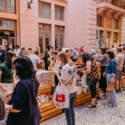 Uma feira ao ar livre acontece em frente a um edif&iacute;cio hist&oacute;rico de fachada rosa e grandes janelas. Diversas pessoas circulam entre bancas de madeira organizadas sobre a cal&ccedil;ada revestida por pedras em padr&atilde;o mosaico. Os expositores apresentam ilustra&ccedil;&otilde;es, objetos artesanais e outros produtos culturais. O movimento intenso de visitantes observando os itens e conversando entre si destaca o car&aacute;ter coletivo e cultural do evento.