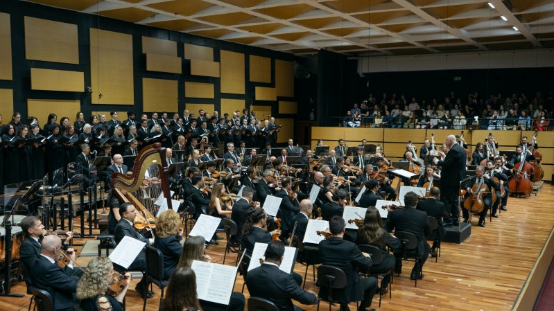 A imagem mostra uma apresenta&ccedil;&atilde;o de uma grande orquestra sinf&ocirc;nica acompanhada por um coro em um audit&oacute;rio. Em primeiro plano, os m&uacute;sicos est&atilde;o sentados em forma&ccedil;&atilde;o semicircular sobre um palco de piso de madeira clara. Eles vestem roupas formais predominantemente pretas e tocam diferentes instrumentos, como violinos, violas, violoncelos, contrabaixos, harpa e instrumentos de sopro. &Agrave; frente da orquestra, sobre um pequeno tablado, o maestro conduz a execu&ccedil;&atilde;o da obra com os bra&ccedil;os erguidos, voltado para os m&uacute;sicos e para o coro. Ao fundo do palco, um coro numeroso ocupa uma plataforma elevada, disposto em v&aacute;rias fileiras. Todos os integrantes tamb&eacute;m usam trajes escuros e est&atilde;o voltados para o maestro. A parede atr&aacute;s do coro &eacute; revestida por pain&eacute;is ac&uacute;sticos geom&eacute;tricos em tons de preto e amarelo, organizados em um padr&atilde;o retangular. &Agrave; direita da imagem, acima do n&iacute;vel do palco, h&aacute; uma plateia sentada em uma varanda, acompanhando a apresenta&ccedil;&atilde;o. A ilumina&ccedil;&atilde;o &eacute; uniforme e destaca o palco, enquanto o teto apresenta uma estrutura quadriculada de pain&eacute;is ac&uacute;sticos.