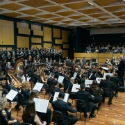 A imagem mostra uma apresenta&ccedil;&atilde;o de uma grande orquestra sinf&ocirc;nica acompanhada por um coro em um audit&oacute;rio. Em primeiro plano, os m&uacute;sicos est&atilde;o sentados em forma&ccedil;&atilde;o semicircular sobre um palco de piso de madeira clara. Eles vestem roupas formais predominantemente pretas e tocam diferentes instrumentos, como violinos, violas, violoncelos, contrabaixos, harpa e instrumentos de sopro. &Agrave; frente da orquestra, sobre um pequeno tablado, o maestro conduz a execu&ccedil;&atilde;o da obra com os bra&ccedil;os erguidos, voltado para os m&uacute;sicos e para o coro. Ao fundo do palco, um coro numeroso ocupa uma plataforma elevada, disposto em v&aacute;rias fileiras. Todos os integrantes tamb&eacute;m usam trajes escuros e est&atilde;o voltados para o maestro. A parede atr&aacute;s do coro &eacute; revestida por pain&eacute;is ac&uacute;sticos geom&eacute;tricos em tons de preto e amarelo, organizados em um padr&atilde;o retangular. &Agrave; direita da imagem, acima do n&iacute;vel do palco, h&aacute; uma plateia sentada em uma varanda, acompanhando a apresenta&ccedil;&atilde;o. A ilumina&ccedil;&atilde;o &eacute; uniforme e destaca o palco, enquanto o teto apresenta uma estrutura quadriculada de pain&eacute;is ac&uacute;sticos.