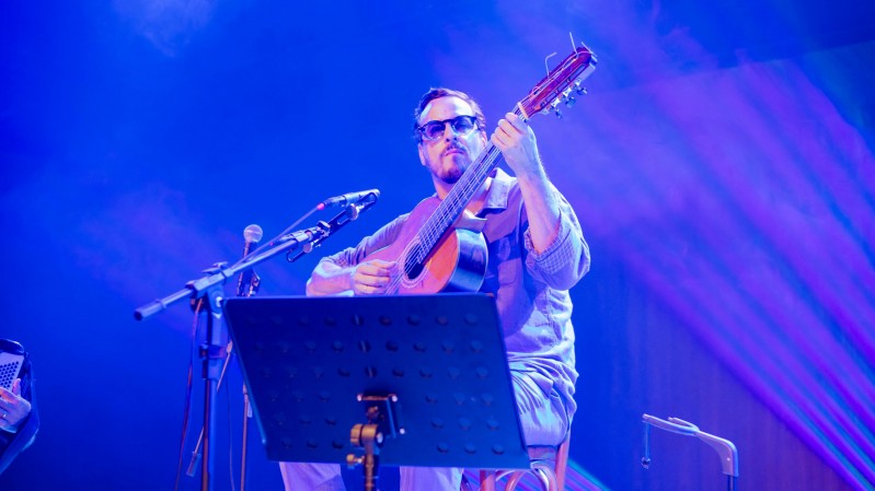 A imagem mostra um m&uacute;sico sentado em um palco, tocando um viol&atilde;o de cordas de nylon durante uma apresenta&ccedil;&atilde;o. Ao centro da imagem est&aacute; o violonista Mathias 7 Cordas, usando &oacute;culos escuros, camisa de mangas compridas dobradas at&eacute; os antebra&ccedil;os e cal&ccedil;a clara. Ele segura o viol&atilde;o junto ao corpo, com a m&atilde;o esquerda posicionada no bra&ccedil;o do instrumento e a direita sobre as cordas, em pleno momento da execu&ccedil;&atilde;o. Seu olhar est&aacute; direcionado para a frente, com express&atilde;o concentrada. &Agrave; frente do m&uacute;sico h&aacute; um pedestal com uma estante de partituras preta, parcialmente cobrindo a parte inferior de seu corpo. &Agrave; esquerda, dois microfones montados em um mesmo suporte est&atilde;o posicionados na dire&ccedil;&atilde;o do viol&atilde;o, indicando a capta&ccedil;&atilde;o do som da apresenta&ccedil;&atilde;o. No canto inferior direito aparece parte de uma cadeira vazia, sugerindo a presen&ccedil;a de outros m&uacute;sicos ou a prepara&ccedil;&atilde;o para uma apresenta&ccedil;&atilde;o em conjunto. 