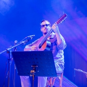A imagem mostra um m&uacute;sico sentado em um palco, tocando um viol&atilde;o de cordas de nylon durante uma apresenta&ccedil;&atilde;o. Ao centro da imagem est&aacute; o violonista Mathias 7 Cordas, usando &oacute;culos escuros, camisa de mangas compridas dobradas at&eacute; os antebra&ccedil;os e cal&ccedil;a clara. Ele segura o viol&atilde;o junto ao corpo, com a m&atilde;o esquerda posicionada no bra&ccedil;o do instrumento e a direita sobre as cordas, em pleno momento da execu&ccedil;&atilde;o. Seu olhar est&aacute; direcionado para a frente, com express&atilde;o concentrada. &Agrave; frente do m&uacute;sico h&aacute; um pedestal com uma estante de partituras preta, parcialmente cobrindo a parte inferior de seu corpo. &Agrave; esquerda, dois microfones montados em um mesmo suporte est&atilde;o posicionados na dire&ccedil;&atilde;o do viol&atilde;o, indicando a capta&ccedil;&atilde;o do som da apresenta&ccedil;&atilde;o. No canto inferior direito aparece parte de uma cadeira vazia, sugerindo a presen&ccedil;a de outros m&uacute;sicos ou a prepara&ccedil;&atilde;o para uma apresenta&ccedil;&atilde;o em conjunto. 