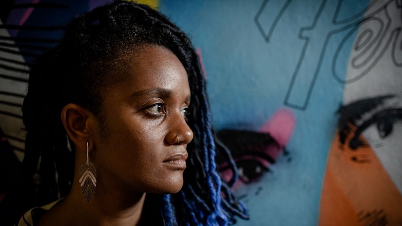 Fotografia em close mostra uma mulher posicionada de perfil, voltada para a direita. Ela &eacute; negra e seus cabelos s&atilde;o longos em tran&ccedil;as com mechas azuladas. Ela usa uma argola no nariz, brinco met&aacute;lico alongado e veste roupa escura. Ao fundo, h&aacute; um mural colorido com predomin&acirc;ncia de tons de azul, mas com detalhes em rosa, preto e laranja, incluindo o desenho parcial de um rosto e de um olho. A ilumina&ccedil;&atilde;o destaca os contornos do rosto e cria contraste entre a figura em primeiro plano e a arte ao fundo.