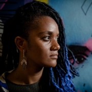 Fotografia em close mostra uma mulher posicionada de perfil, voltada para a direita. Ela &eacute; negra e seus cabelos s&atilde;o longos em tran&ccedil;as com mechas azuladas. Ela usa uma argola no nariz, brinco met&aacute;lico alongado e veste roupa escura. Ao fundo, h&aacute; um mural colorido com predomin&acirc;ncia de tons de azul, mas com detalhes em rosa, preto e laranja, incluindo o desenho parcial de um rosto e de um olho. A ilumina&ccedil;&atilde;o destaca os contornos do rosto e cria contraste entre a figura em primeiro plano e a arte ao fundo.