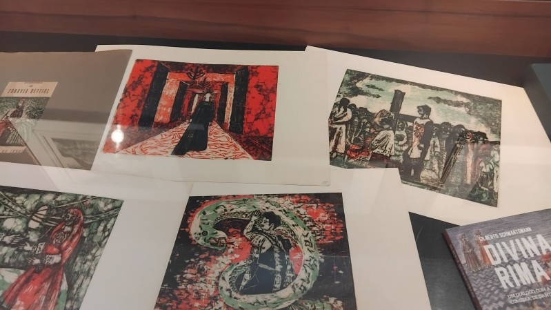 Fotografia de uma vitrine expositiva vista de cima, contendo gravuras e publica&ccedil;&otilde;es organizadas sobre uma superf&iacute;cie escura. As obras apresentam cenas figurativas em cores intensas, com predomin&acirc;ncia de vermelho, preto, verde e branco. Entre as imagens, aparecem uma figura em um corredor vermelho, personagens agrupados em uma cena ao ar livre, uma pessoa envolvida por uma serpente e uma figura vestida de vermelho em meio &agrave; vegeta&ccedil;&atilde;o. &Agrave; direita, h&aacute; um livro com o t&iacute;tulo &ldquo;Divina Rima&rdquo;, enquanto no canto superior esquerdo aparece parcialmente outro volume apoiado dentro da vitrine. Reflexos da ilumina&ccedil;&atilde;o sobre o vidro evidenciam o car&aacute;ter museol&oacute;gico da montagem.