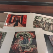 Fotografia de uma vitrine expositiva vista de cima, contendo gravuras e publica&ccedil;&otilde;es organizadas sobre uma superf&iacute;cie escura. As obras apresentam cenas figurativas em cores intensas, com predomin&acirc;ncia de vermelho, preto, verde e branco. Entre as imagens, aparecem uma figura em um corredor vermelho, personagens agrupados em uma cena ao ar livre, uma pessoa envolvida por uma serpente e uma figura vestida de vermelho em meio &agrave; vegeta&ccedil;&atilde;o. &Agrave; direita, h&aacute; um livro com o t&iacute;tulo &ldquo;Divina Rima&rdquo;, enquanto no canto superior esquerdo aparece parcialmente outro volume apoiado dentro da vitrine. Reflexos da ilumina&ccedil;&atilde;o sobre o vidro evidenciam o car&aacute;ter museol&oacute;gico da montagem.
