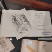 Fotografia de uma vitrine expositiva com livros raros e gravuras abertas para apreciação do público. No centro, uma grande página exibe o título em francês “Romances sans paroles, Chansons pour elle, Odes en son honneur”, acompanhado da indicação de gravuras de Mariette Lydis. Sobre as páginas está apoiada uma ilustração em preto e branco retratando uma figura sentada. Ao redor, outros livros e documentos permanecem abertos, revelando desenhos delicados e páginas antigas. Pequenas legendas identificam as peças expostas, enquanto o reflexo da iluminação sobre o vidro reforça o ambiente de preservação e exposição do acervo.