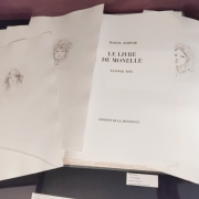 Fotografia de uma vitrine expositiva vista de cima, com páginas soltas, gravuras e publicações dispostas sobre uma superfície escura. No centro, uma grande página aberta traz o título em francês “Le Livre de Monelle”, acompanhado dos nomes Marcel Schwob e Leonor Fini. Ao redor, aparecem delicados desenhos em preto e branco de rostos e figuras humanas impressos em folhas avulsas. Pequenas legendas identificam as peças expostas.