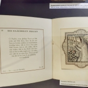 Fotografia de um livro aberto em uma vitrine de exposição. Na página da esquerda há um texto em alemão com o título “Die Gläubigen Frauen”, organizado em blocos tipográficos. Na página da direita, uma ilustração colorida mostra uma figura envolta por vestes ornamentadas, cercada por elementos decorativos e florais em estilo inspirado na arte oriental e no art nouveau. Acima do livro, uma legendagem apresenta informações sobre a ilustradora Élisabeth Sonrel. A peça está protegida por vidro, e a iluminação suave destaca os detalhes da publicação histórica.