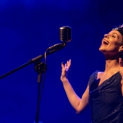 A imagem mostra uma pessoa em um palco, posicionada &agrave; direita da composi&ccedil;&atilde;o, diante de um microfone de estilo retr&ocirc; montado em um pedestal. A pessoa est&aacute; com os bra&ccedil;os erguidos e as m&atilde;os abertas pr&oacute;ximas ao rosto, em uma postura que sugere uma apresenta&ccedil;&atilde;o art&iacute;stica ou musical. O ambiente &eacute; iluminado por uma intensa luz azul, que domina tanto o fundo quanto a ilumina&ccedil;&atilde;o da cena. O fundo &eacute; liso e escuro, sem elementos vis&iacute;veis al&eacute;m do efeito da ilumina&ccedil;&atilde;o c&ecirc;nica. O microfone est&aacute; inclinado em dire&ccedil;&atilde;o &agrave; pessoa e ocupa a regi&atilde;o central da imagem. A pessoa veste uma roupa escura e brilhante, sem mangas, com textura que reflete a luz do palco. Tamb&eacute;m &eacute; poss&iacute;vel observar um acess&oacute;rio na cabe&ccedil;a com detalhes claros que se destacam sob a ilumina&ccedil;&atilde;o azul. N&atilde;o h&aacute; textos, logotipos ou outros objetos vis&iacute;veis na imagem.