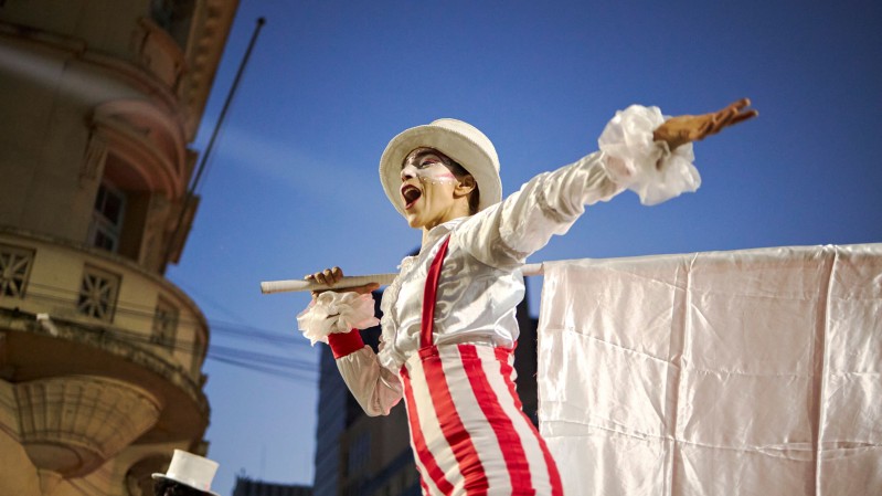 Fotografia realizada ao ar livre ao entardecer mostra uma pessoa caracterizada para uma apresenta&ccedil;&atilde;o art&iacute;stica em espa&ccedil;o urbano. A pessoa veste roupa branca com detalhes vermelhos, incluindo cal&ccedil;as listradas, suspens&oacute;rios e chap&eacute;u claro. Com um dos bra&ccedil;os estendido para a frente, segura sobre os ombros uma haste da qual pende um grande tecido branco. A cena &eacute; registrada em &acirc;ngulo baixo, destacando o c&eacute;u azul ao fundo e parte da fachada de um pr&eacute;dio hist&oacute;rico com sacada e elementos ornamentais. A ilumina&ccedil;&atilde;o valoriza o contraste entre o figurino claro e o ambiente urbano ao redor.